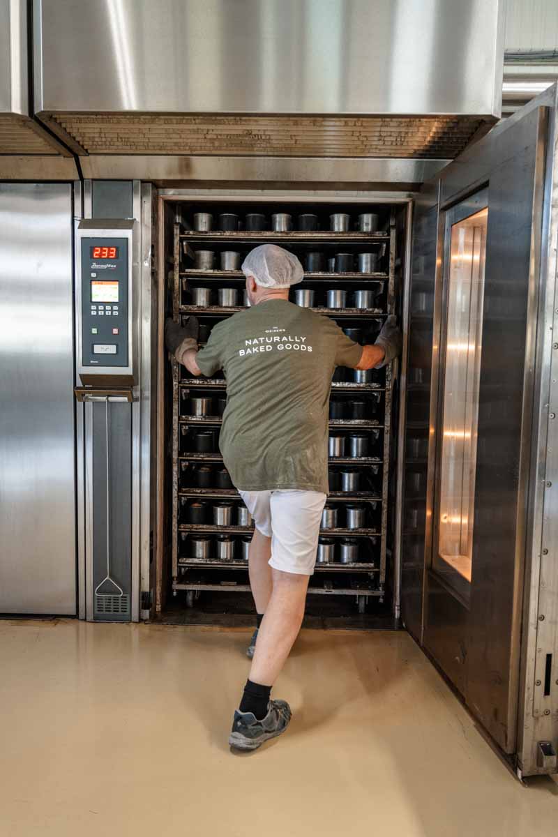 Christian Deiser schiebt gerade ein Backblech mit Dosenbroten in einen Industriebackofen. Im Ofen stehen schon sehr viele Brote. Der Bäcker ist von hinten zu sehen. Er trägt ein grünes T-Shirt mit dem Claim "Naturally baked goods", eine Bäckermütze, eine kurze weiße Hose und schwarze Schuhe.