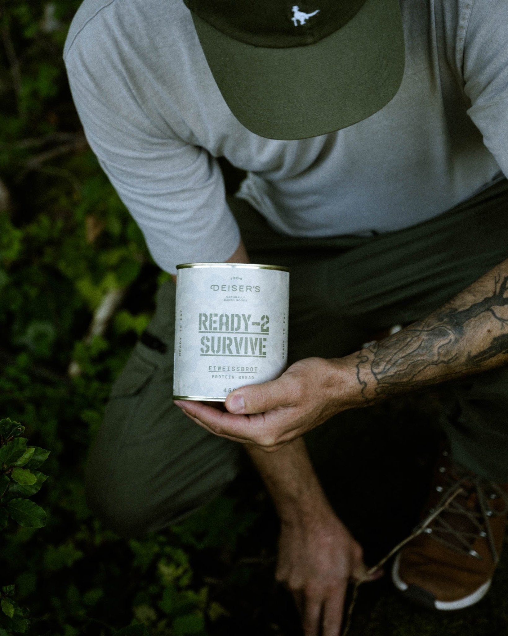 Ein Mann im Outdoor-Trekking-Outfit kniet im Wald und hält eine Dose Deiser's Ready-2-Survive Eiweißbrot in der Hand.