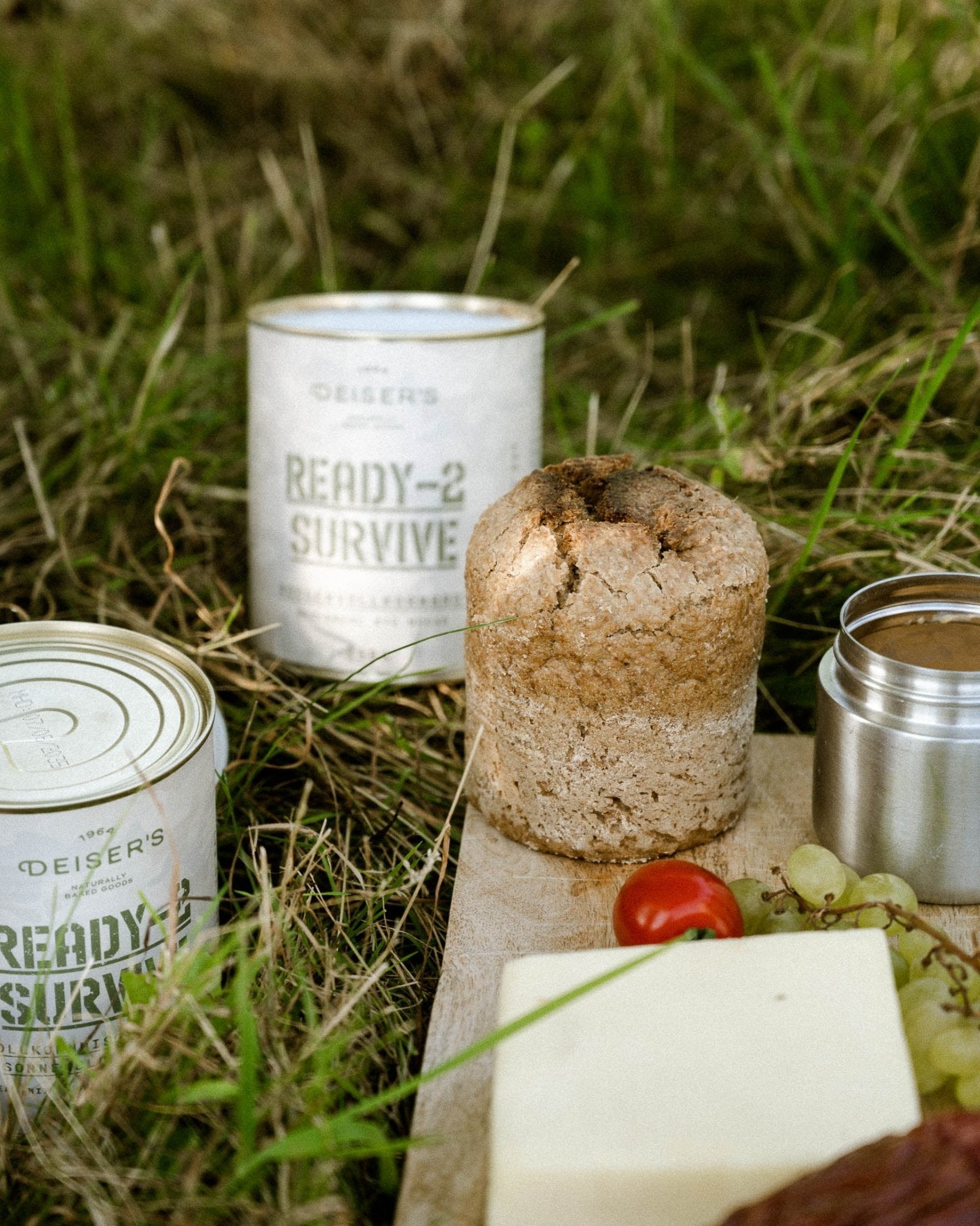 Eine offene und eine geschlossen Dose Deiser's Dosembrot stehen in der Wiese, davor ein Holzbrett mit einem Dosenbrot, Gemüse und Käse.