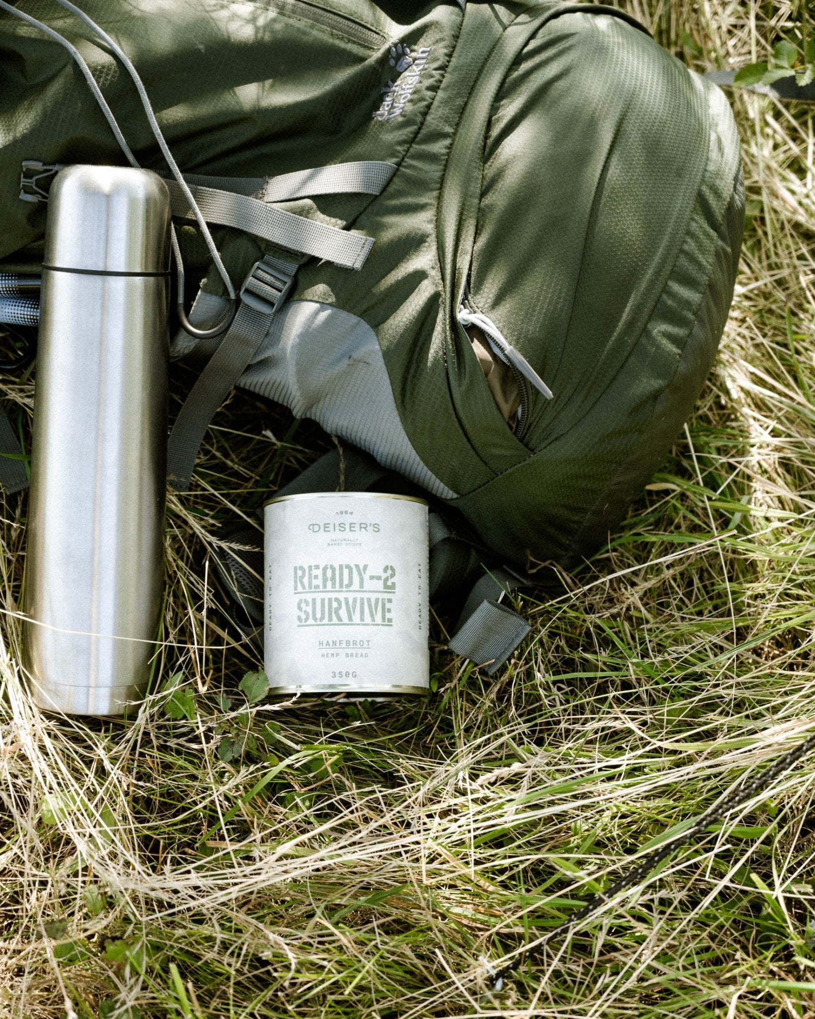 Eine Dose Deiser's Ready-2-Survive Hanfbrot liegt in der Wiese neben einem Rücksack und einer Thermoskanne.