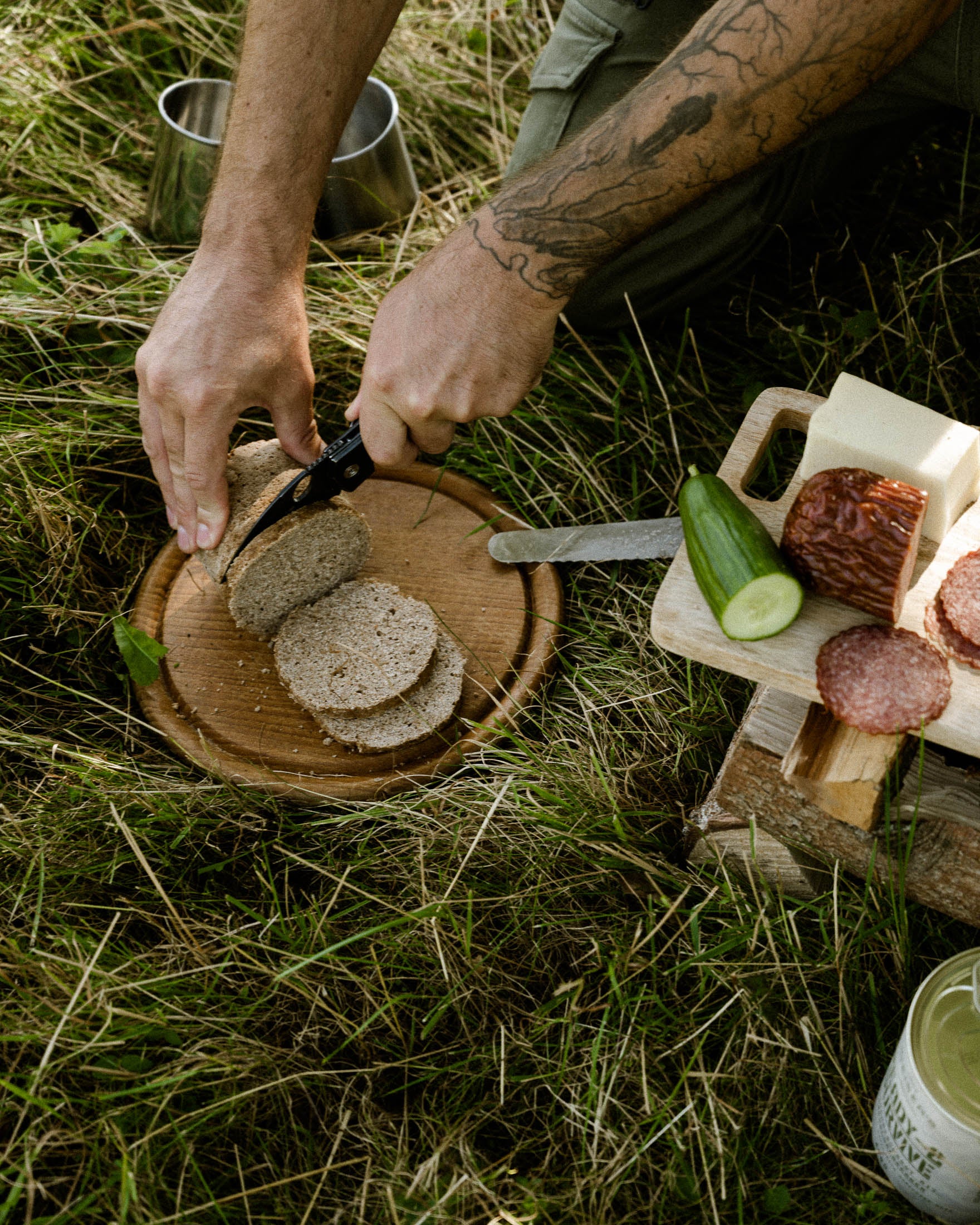 Mit einem Taschenmesser schneidet ein Mann Brotscheiben des Ready-2-Survive Dosenbrots von Deiser's. Daneben in der Wiese liegen Wurst, Käse und Gurke auf einem Holzbrett.