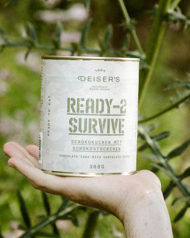 Eine Hand hält eine Dose Ready-2-Survive Schokokuchen mit Schokostückchen. Im Hintergrund sieht man eine grüne Pflanze. 